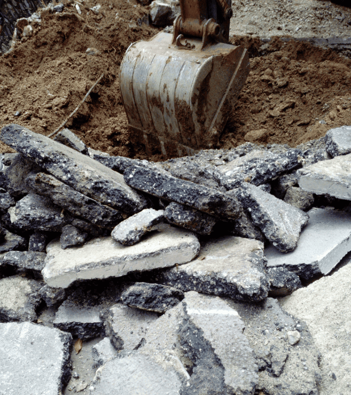 Restos de asfalto y hormigón acumulados mientras una excavadora retira tierra en una obra para su posterior recogida y reciclaje.